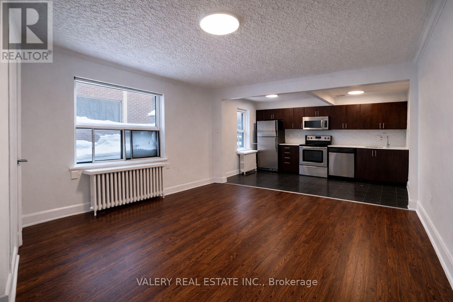 A room with wood flooring and a kitchen area in the background.