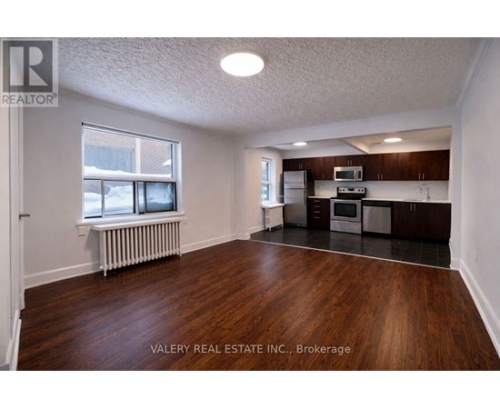 A room with wood flooring and a kitchen area in the background.