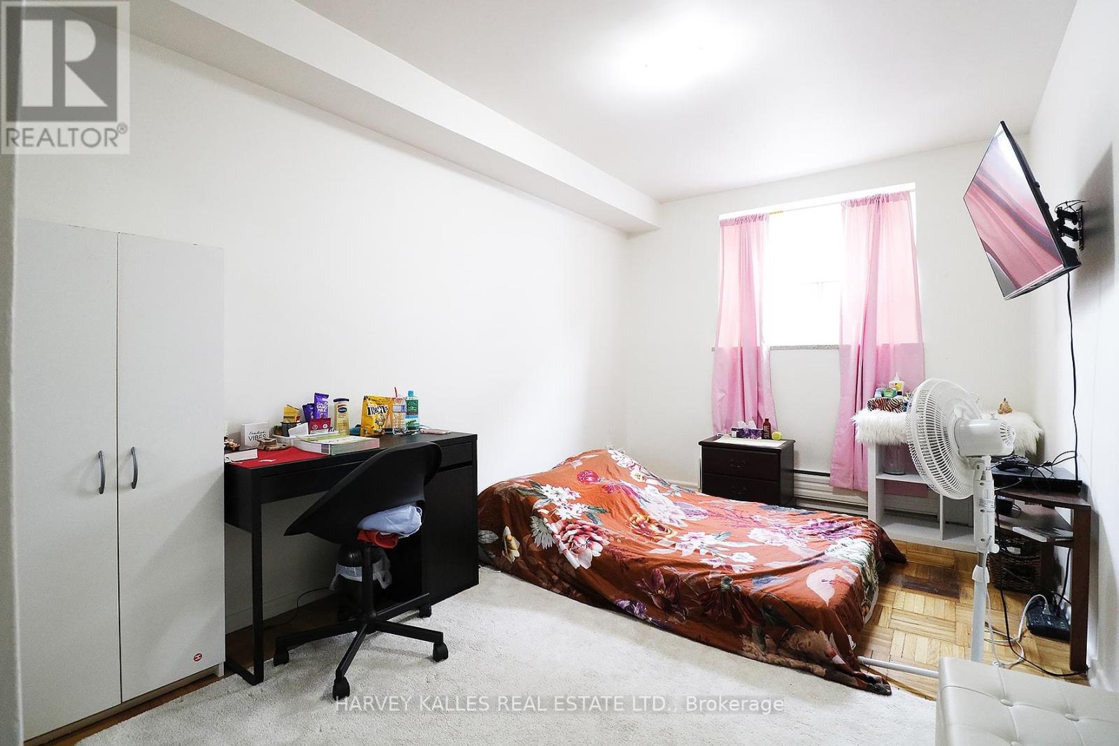 A bedroom with a bed covered in a pink bedspread, a desk with a chair, and a fan.