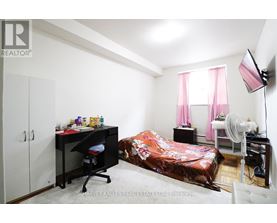 A bedroom with a bed covered in a pink bedspread, a desk with a chair, and a fan.