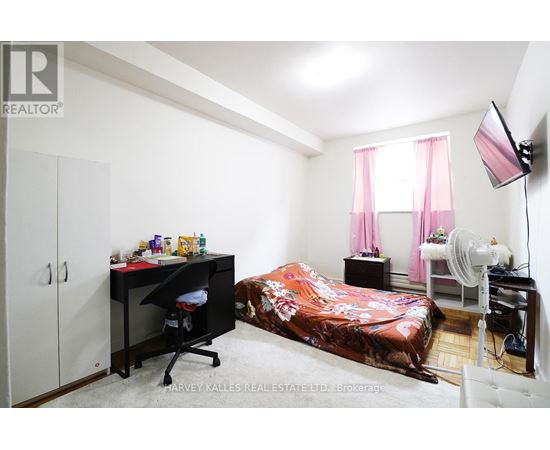 A bedroom with a bed covered in a pink bedspread, a desk with a chair, and a fan.