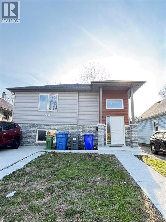 A house with a red car parked in front and three trash bins in front of the house.