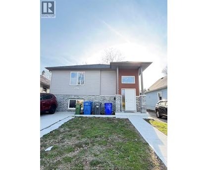 A house with a red car parked in front and three trash bins in front of the house.