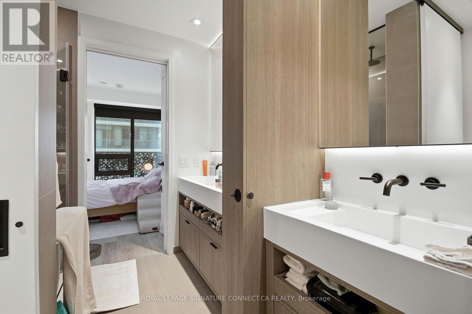 A bathroom with a sink, mirror, and a bedroom with a bed and window in the background.