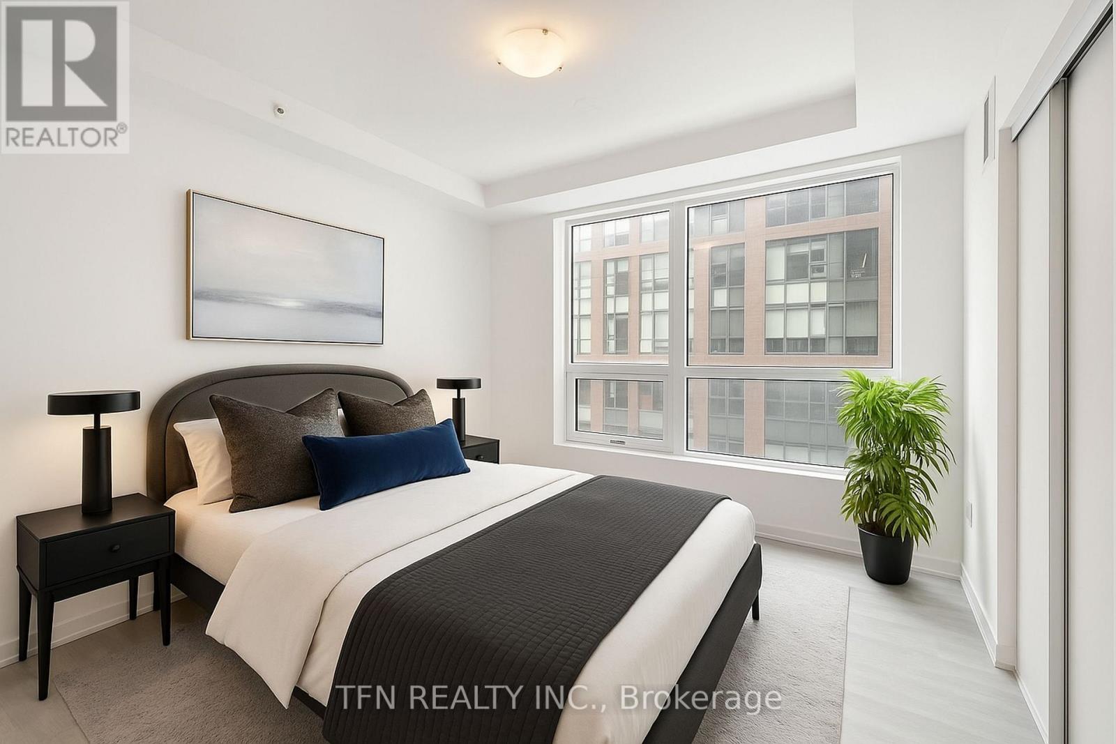 A bedroom with a large bed, a nightstand with a lamp, a potted plant, and a window with a view of a building.