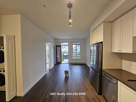 A cat is sitting on a wooden floor in a kitchen with white cabinets and a black fridge.