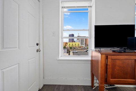 A room with a white door, a window with a view of a cityscape, and a wooden desk with a computer on it.