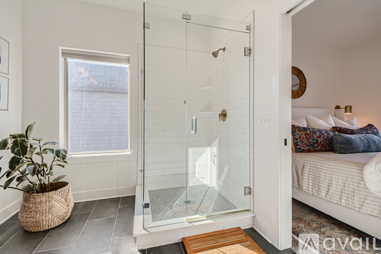A bathroom with a glass shower door and a bedroom with a bed and a window.