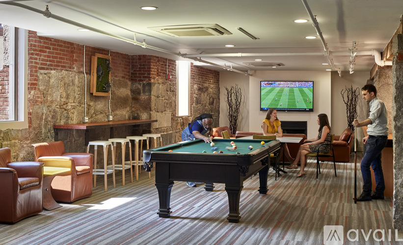 A group of people playing pool and watching a soccer game on television in a room with a brick wall.