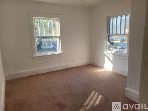 A room with a carpeted floor and two windows letting in sunlight.