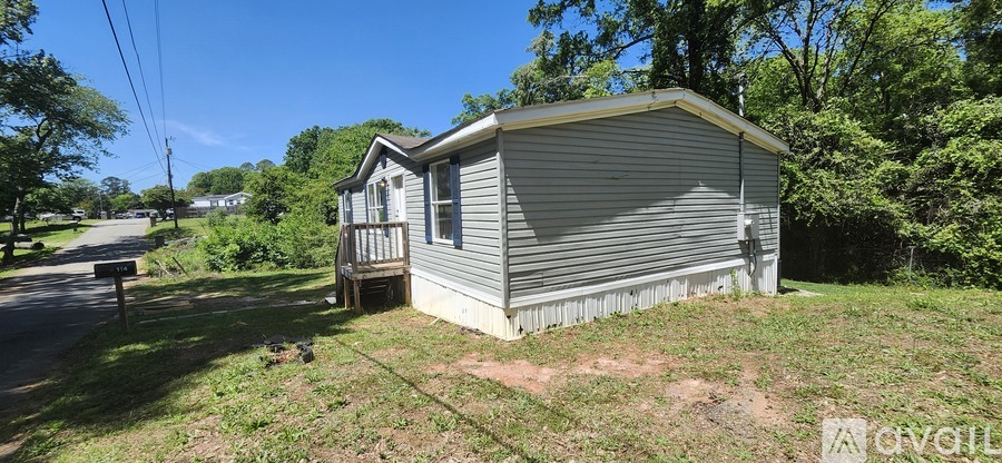 A small house with a grey roof and a porch is surrounded by greenery.