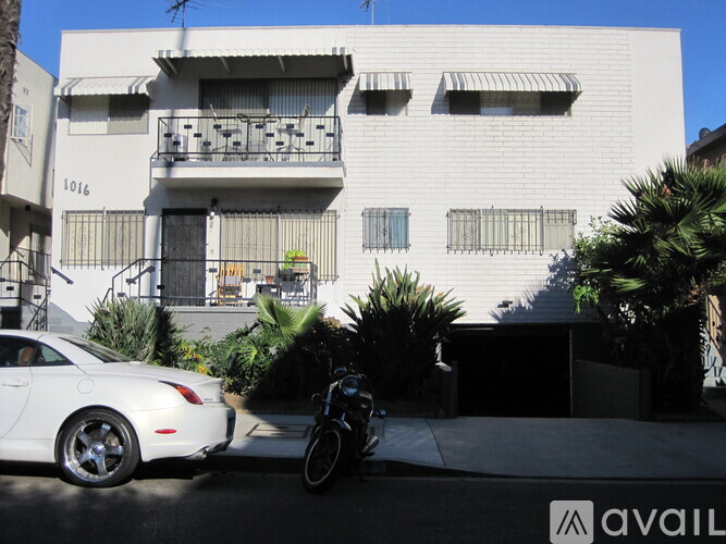 A white car is parked in front of a white building with a balcony.