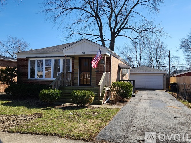 A house with a flag on the front porch is surrounded by a driveway and a tree.