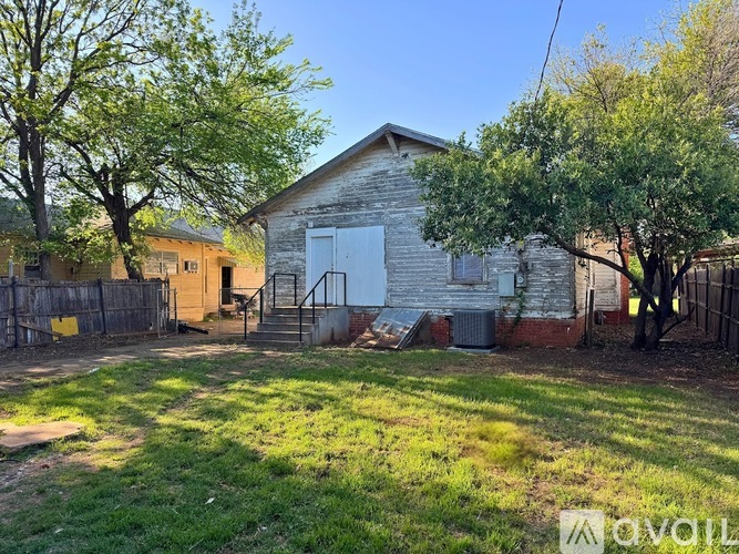 A small, old building with a white door is surrounded by a fence and trees.