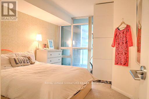 A bedroom with a white bed, a nightstand with a lamp, a window with a view of the outside, and a red dress hanging on the door.