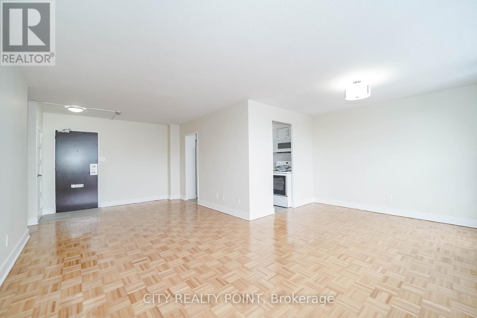 A spacious room with wooden flooring and white walls, featuring a refrigerator and a small kitchenette.