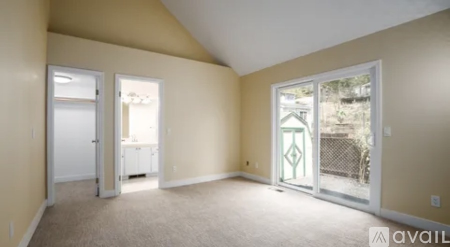 A room with beige walls and carpet, two doors, and a window with a view of a fence and trees outside.