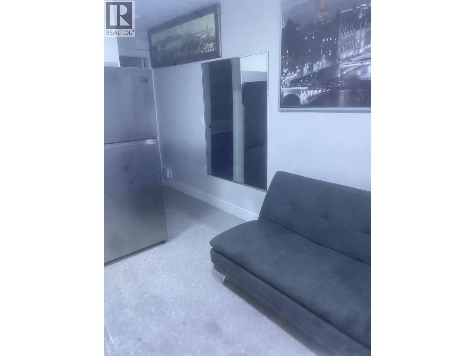 A grey couch sits in a room with a fridge and a picture on the wall.