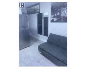 A grey couch sits in a room with a fridge and a picture on the wall.