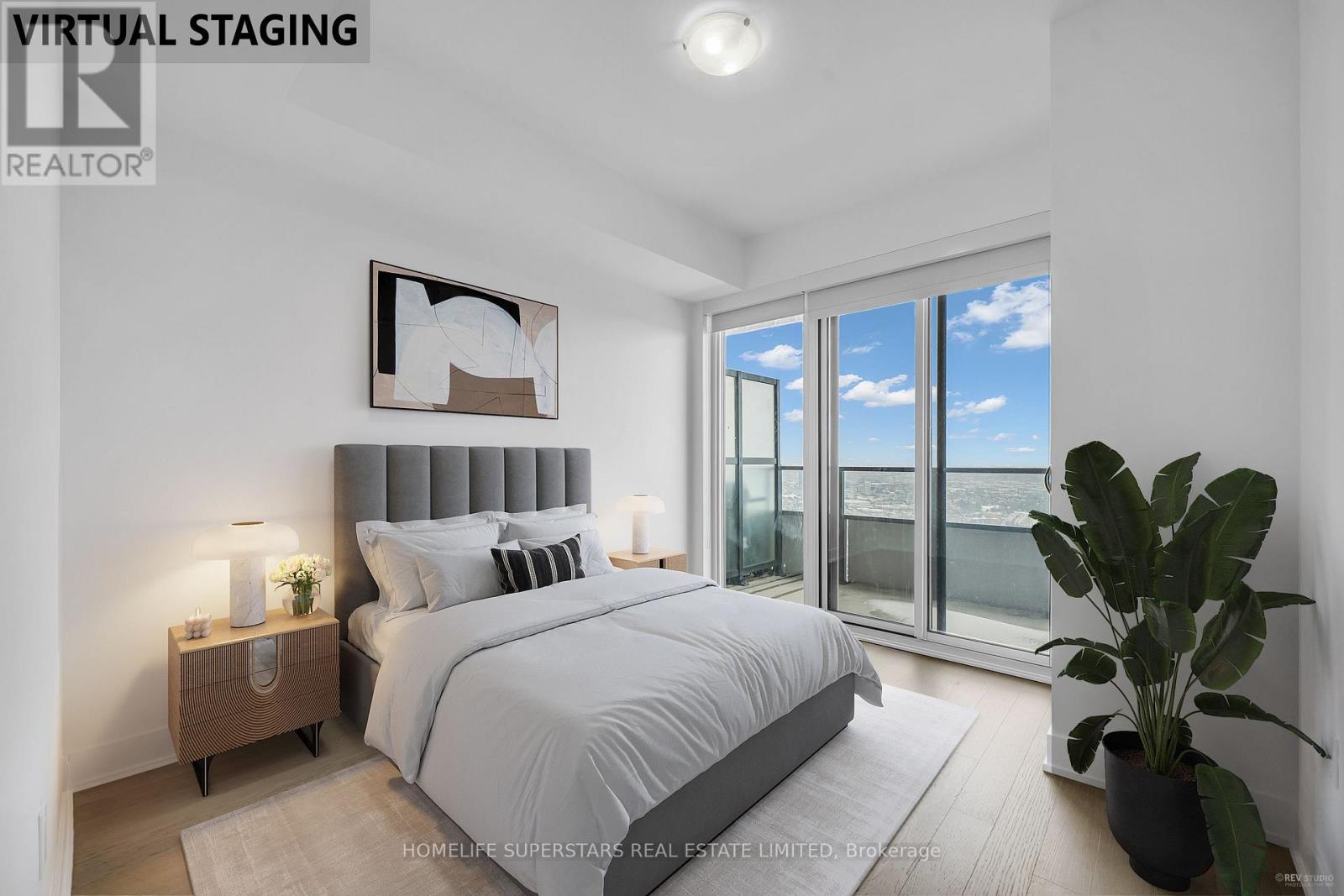 A bedroom with a large bed, a nightstand with a lamp, a potted plant, and a framed picture on the wall.