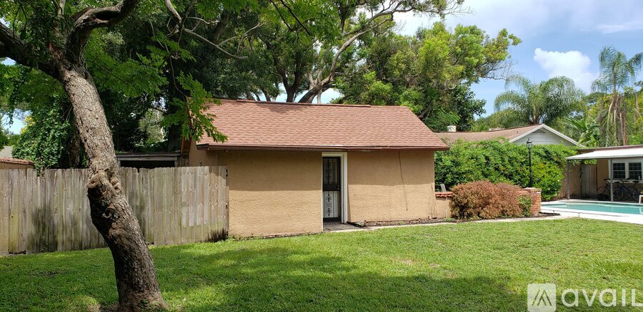 A small house with a brown roof and tan walls is surrounded by a fence and trees.