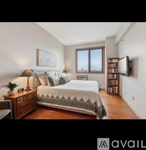 A bedroom with a large bed, a nightstand with a lamp, a bookshelf, and a window with a view.