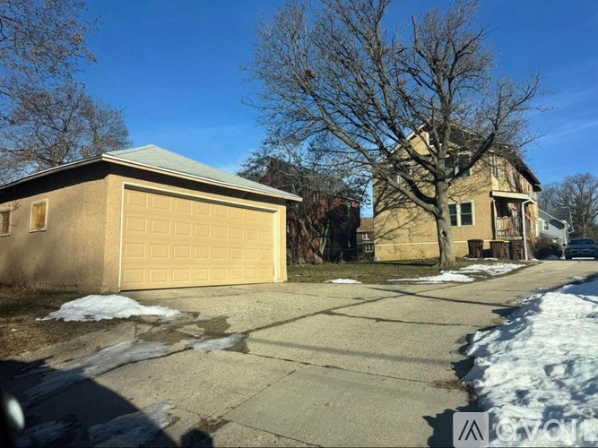 A small tan building with a garage door is in front of a bare tree and a house.