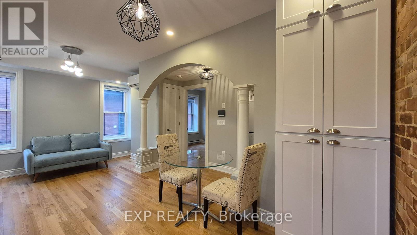 A real estate listing photo of a living room with a grey couch, a glass table, and a brick wall.