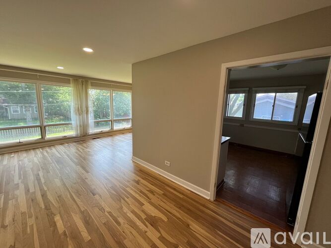 A room with wooden flooring and a large window with a view of a fence and trees.