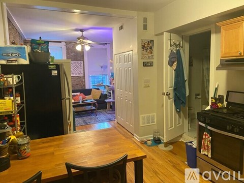 A kitchen with a wooden table and chairs in the foreground and a living room with a couch and a fan in the background.
