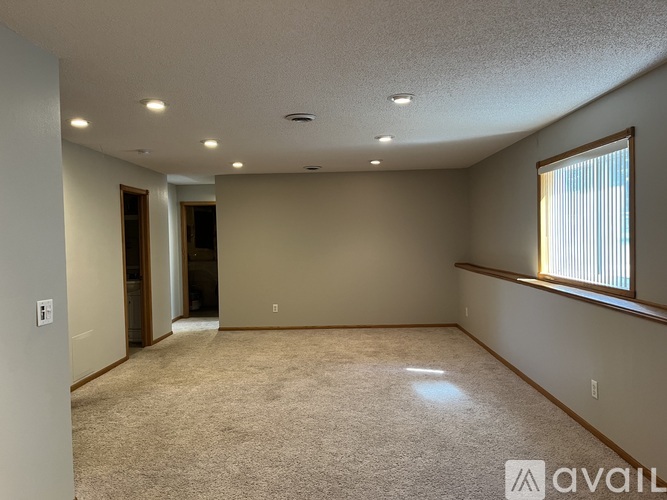 A spacious room with carpeted floor, a window with blinds, and a door leading to another room.