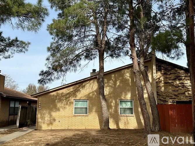 A house with a brown brick exterior is surrounded by trees and has a fence in front.