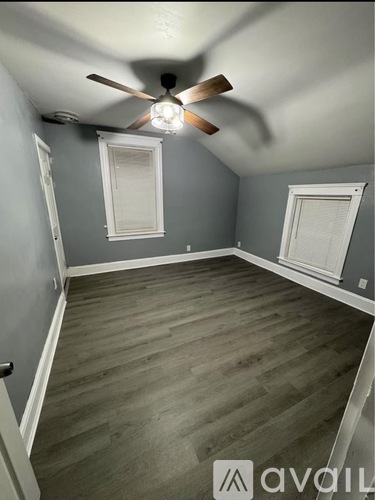 A room with a ceiling fan and light, wooden flooring, and two windows with white frames.