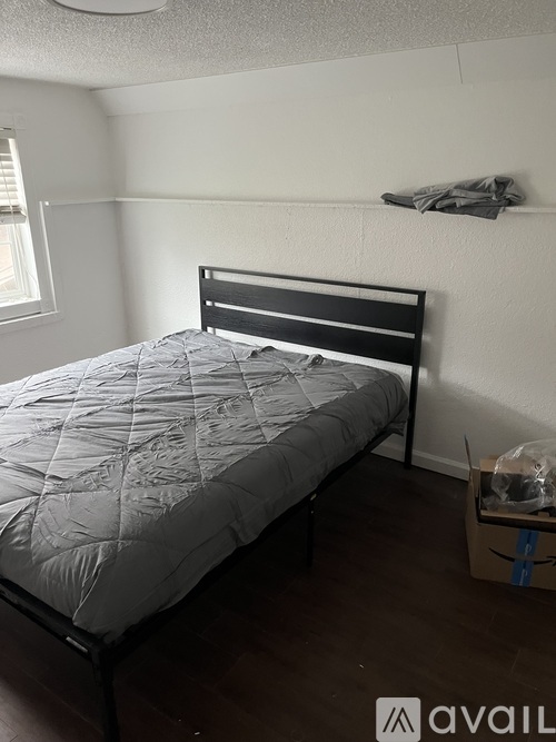 A bed with a grey comforter is in a room with a white wall and a window.
