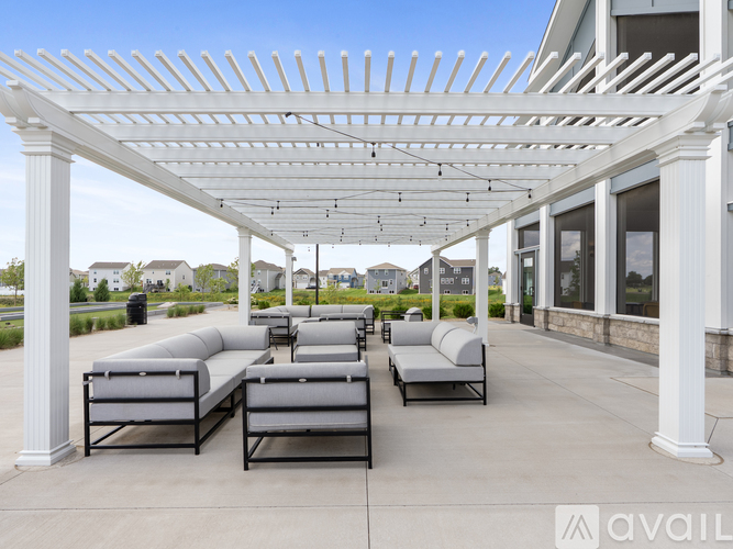A white pergola with grey cushioned seating is in front of a building.