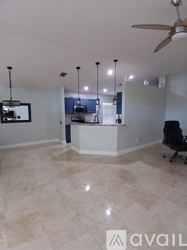 A room with a fan, a chair and a kitchen area in the background.