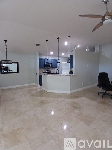 A room with a fan, a chair and a kitchen area in the background.