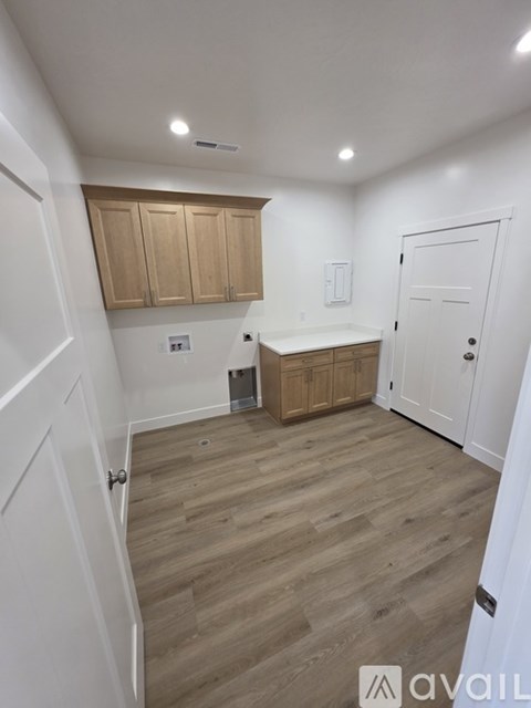 A room with wooden flooring and white walls, featuring a kitchenette with wooden cabinets and a white door.