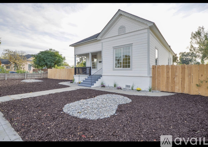 A house with a white exterior and a grey roof is surrounded by a wooden fence and has a gravel garden area in front.