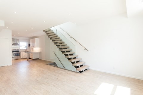 A staircase with wooden steps and a metal handrail is in the middle of a spacious room with a kitchen in the background.