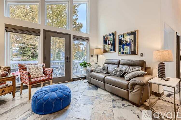 A living room with a brown leather couch, a blue ottoman, and a wooden table with a red and white patterned chair.