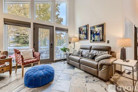 A living room with a brown leather couch, a blue ottoman, and a wooden table with a red and white patterned chair.