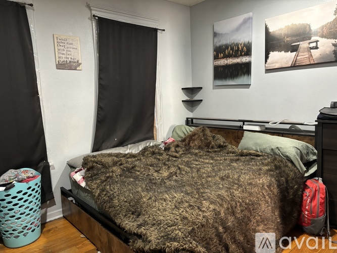A bedroom with a bed covered in a brown furry blanket, a blue laundry basket, a red backpack, and two pictures on the wall.