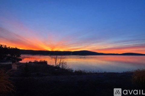 A serene landscape with a lake, a tree, and a sky painted with hues of orange and blue.
