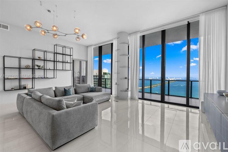 A modern living room with a large grey sofa and a view of the sea through the windows.