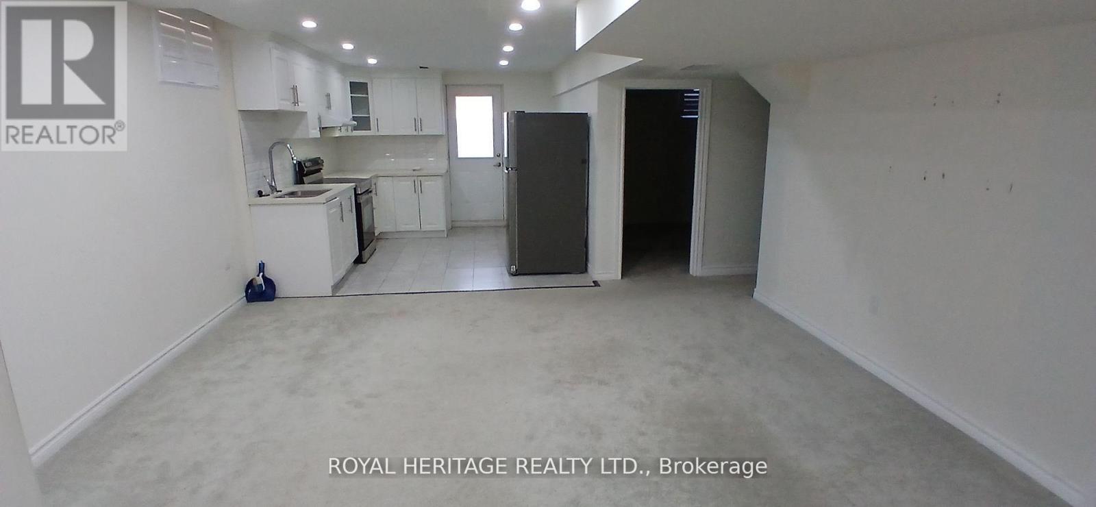 A spacious, empty room with white walls and a kitchen area in the background.