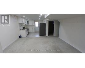 A spacious, empty room with white walls and a kitchen area in the background.