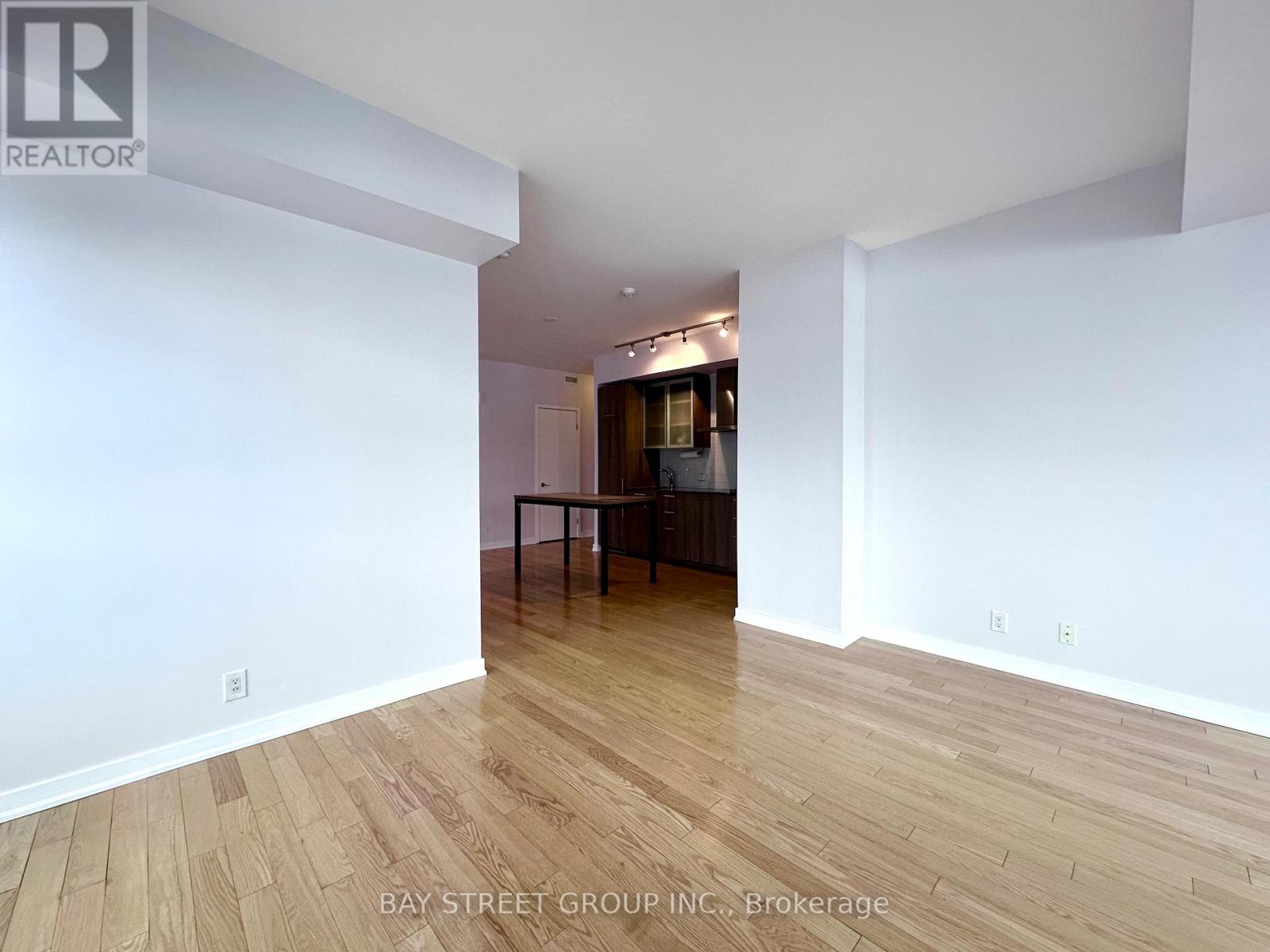 A spacious room with wooden flooring and white walls, with a dining area in the background.