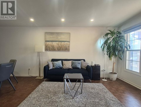 A living room with a black couch, a grey rug, a wooden floor, a potted plant, and a painting on the wall.