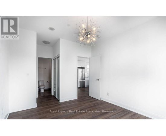A spacious room with a wooden floor and white walls, featuring a large chandelier and a doorway leading to another room.